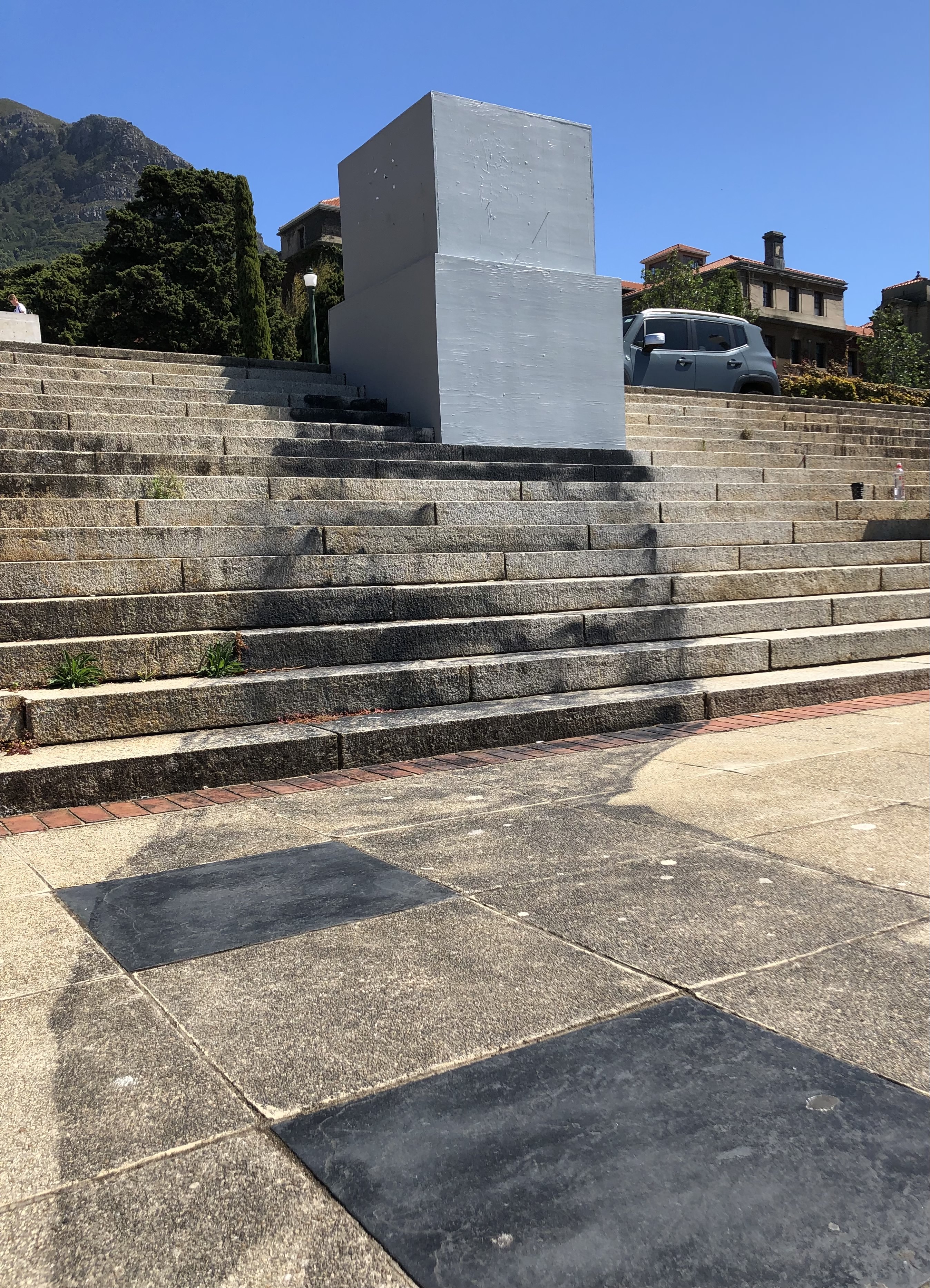 A grey, boxy plinth spans the rise of several steps at the top of an outdoor staircase. A shadowy form pours from the plinth and seeps across the pavers in front of us. It looks like the concrete has been darkened over time.