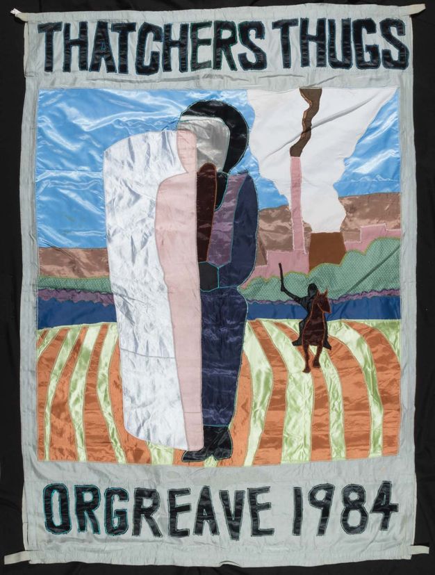 Textile. Created with patches of simmering fabric, an abstracted person stands in a field, close to us. Another riding a horse in the distance holds up a stick or a weapon. A narrow smokestack and a wide, nuclear smokestack are in the background.