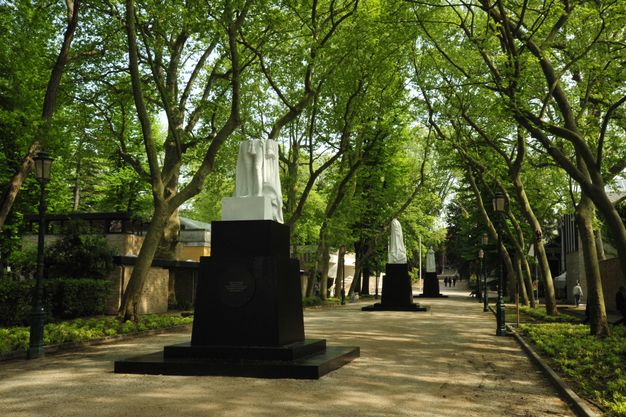 Public art. Three black plinths in a tree-covered lane hold what appear to be fragments of white statues. The statue closest to us seems broken off above the hips.