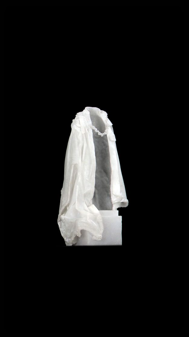 Video still. A white cloak hangs as if over someone’s shoulders, but is empty. The opening is angled to our right against a black background.