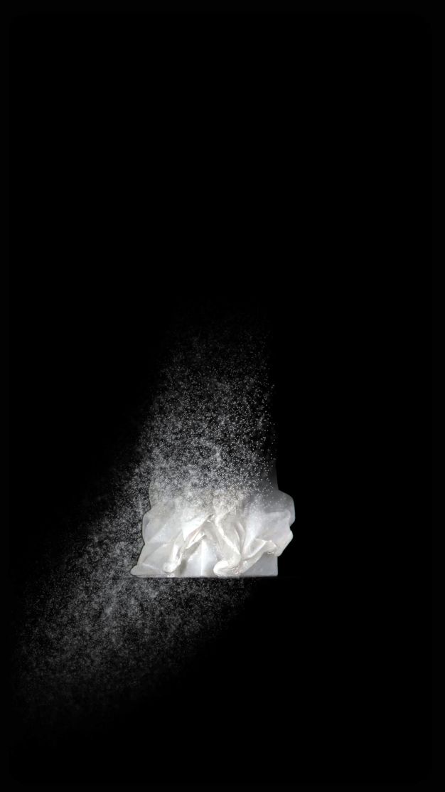Video still. Most of a white cloak has dissolved, leaving a spray of white specks and drapery folded around a square base against a black background.