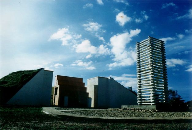 Architecture. Three blocky, angular structures sit in a row to our left, across a plaza. To our right, a square tower appears to be made from boards, like a structure made with popsicle sticks but on a monumental scale.