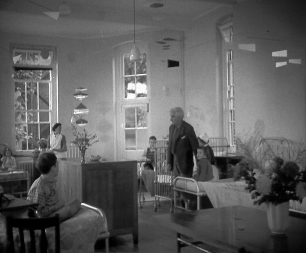 Black and white photograph. Children's ward with five young children seated up in bed in a room with high ceilings, tall windows, and bouquets in vases. A senior person wearing a suit jacket and pants appears to walk across the center of the room with two nurses in uniform attending to the children in the background.